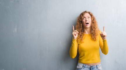 Young redhead woman over grey grunge wall amazed and surprised looking up and pointing with fingers and raised arms.