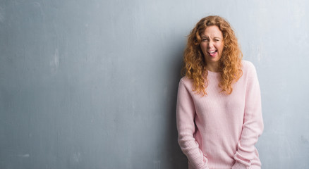 Young redhead woman over grey grunge wall wearing pink sweater sticking tongue out happy with funny expression. Emotion concept.