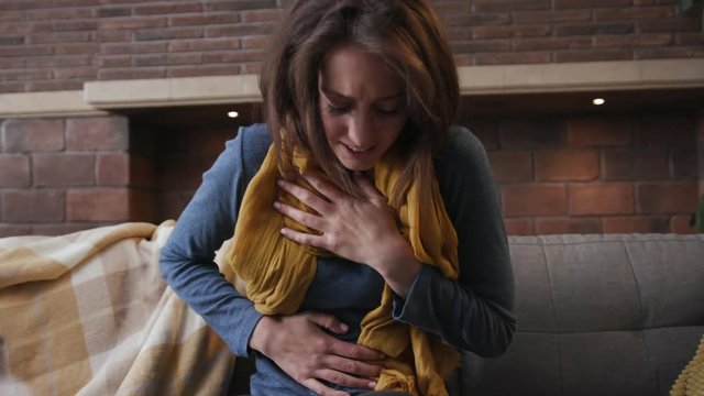 young woman stomach ache sitting in home on sofa feel ill unhappy ache adult female symptom health unhealthy young infection pills medicine portrait screaming pain