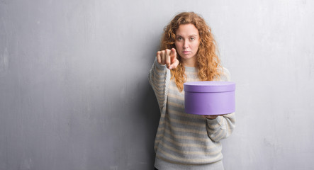 Young redhead woman over grey grunge wall holding box pointing with finger to the camera and to...
