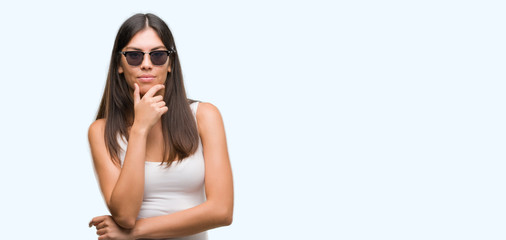 Young beautiful hispanic wearing sunglasses looking confident at the camera with smile with crossed arms and hand raised on chin. Thinking positive.
