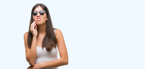Fototapeta premium Young beautiful hispanic wearing sunglasses touching mouth with hand with painful expression because of toothache or dental illness on teeth. Dentist concept.