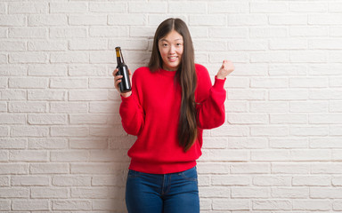 Young Chinese woman over brick wall drinking beer screaming proud and celebrating victory and success very excited, cheering emotion