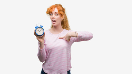 Young redhead woman holding alarm clock with surprise face pointing finger to himself