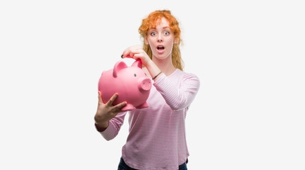 Young redhead woman holding piggy bank scared in shock with a surprise face, afraid and excited with fear expression