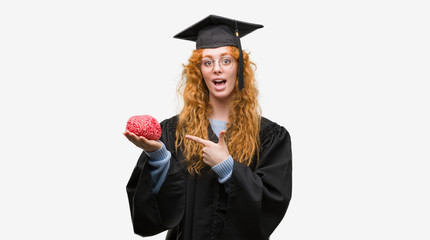 Young redhead student woman wearing graduated uniform holding brain very happy pointing with hand and finger