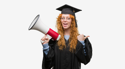 Young redhead student woman wearing graduated uniform holding megaphone pointing and showing with...