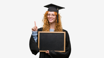 Young redhead student woman wearing graduated uniform holding blackboard surprised with an idea or...