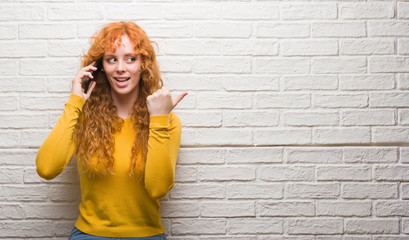 Young redhead woman standing over brick wall talking on the phone pointing and showing with thumb...