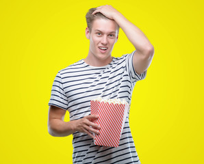 Young handsome blond man eating popcorn stressed with hand on head, shocked with shame and surprise face, angry and frustrated. Fear and upset for mistake.