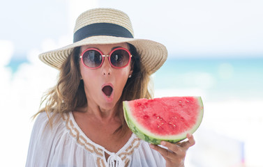 Middle age brunette woman by the sea eating watermelon scared in shock with a surprise face, afraid and excited with fear expression