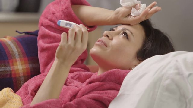 Woman sick at home with cold and flu holding thermometer