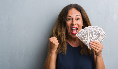 Middle age hispanic woman standing over grey grunge wall holding dollars screaming proud and...