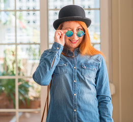 Stylish redhead woman wearing bowler hat and sunglasses winking looking at the camera with sexy...