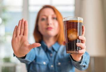 Redhead woman holding soda refreshment with open hand doing stop sign with serious and confident...