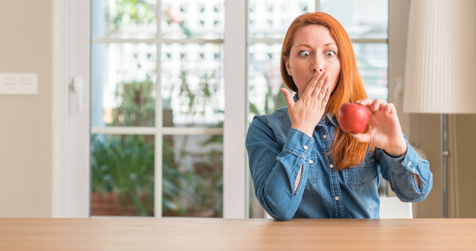 Redhead woman holding red apple at home cover mouth with hand shocked with shame for mistake, expression of fear, scared in silence, secret concept