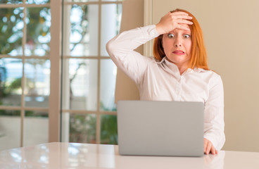 Redhead woman using computer laptop at home stressed with hand on head, shocked with shame and...