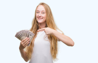 Blonde teenager woman holding dollars very happy pointing with hand and finger