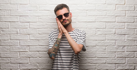 Young adult man wearing sunglasses standing over white brick wall sleeping tired dreaming and posing with hands together while smiling with closed eyes.