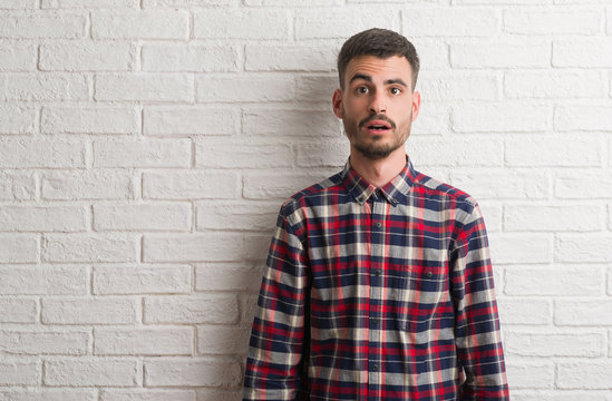 Young adult man standing over white brick wall afraid and shocked with surprise expression, fear and excited face.