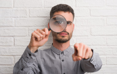 Young adult man over brick wall using magnifying glass pointing with finger to the camera and to...