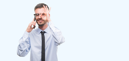 Young business adult man talking on the phone with happy face smiling doing ok sign with hand on eye looking through fingers