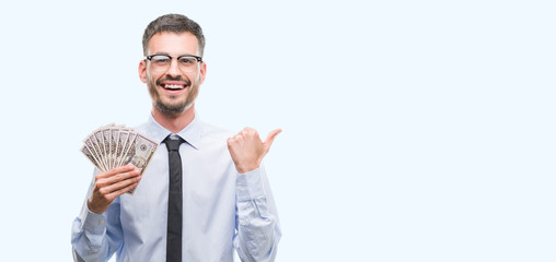 Young hipster business man holding dollars pointing and showing with thumb up to the side with...