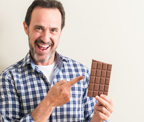 Senior man eating chocolate bar very happy pointing with hand and finger