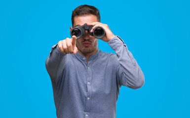 Handsome young man looking through binoculars pointing with finger to the camera and to you, hand sign, positive and confident gesture from the front