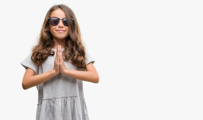 Brunette hispanic girl wearing sunglasses praying with hands together asking for forgiveness smiling confident.