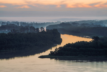 Early blue sky reflected in river water. Riverbank with forest under predawn sky. Cloudy sunrise sky on background. Fog hid trees on island. Colorful morning atmospheric landscape of majestic nature.