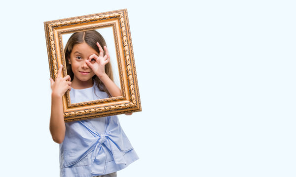 Brunette hispanic girl holding vintage art frame with happy face smiling doing ok sign with hand on eye looking through fingers