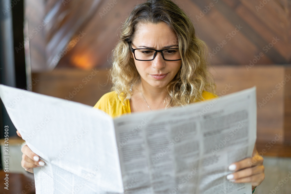 Wall mural frowning young woman reading job ads in newspaper. displeased girl reading article in local newspape