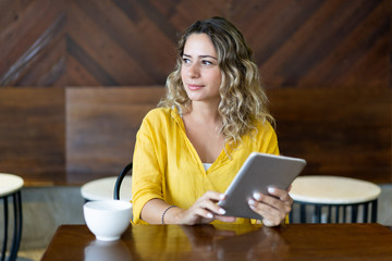 Pensive pretty young woman sitting at cafe table with tablet. Beautiful smiling girl reading book...