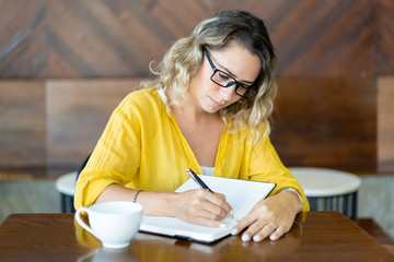 Pretty young woman drinking coffee and writing plans for day. Concentrated girl filling her diary...