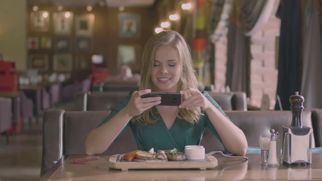 Happy caucasian blonde woman smiling and taking a food picture on the cellphone in cafe. Beautiful lady sitting on the sofa and photographed dish. Luxury grill restaurant and steak on the wooden table