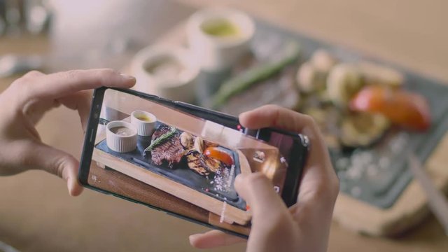 The smartphone screen and food photo concept. Woman's hands making a few picture of delicious steak with vegetables at wooden tray in the grill restaurant from the top, pushing on the touchscreen