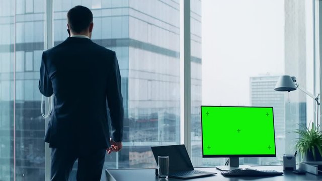Confident Businessman in His Office Making Phone Call, Personal Computer on His Desk Shows Green Chroma Key Screen. In the Window Panoramic Big City View.