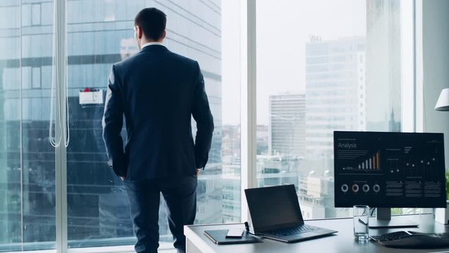 Confident Businessman in a Suit Contemplating Business Deal in His Office, Stands up from His Desk and Walks to a Window to Think. Window Has Panoramic View on Big City Business District.