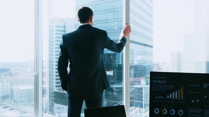 Confident Businessman in a Suit Walking Through His Office and Looking out of the Window Thoughtfully. Stylish Modern Business Office with Personal Computer and Big City View.