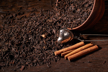 Dried black tea in clay bowl with sticks of cinnamon and a tea strainer isolated on black background