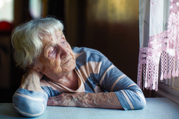 An elderly woman sits sadly near the window.