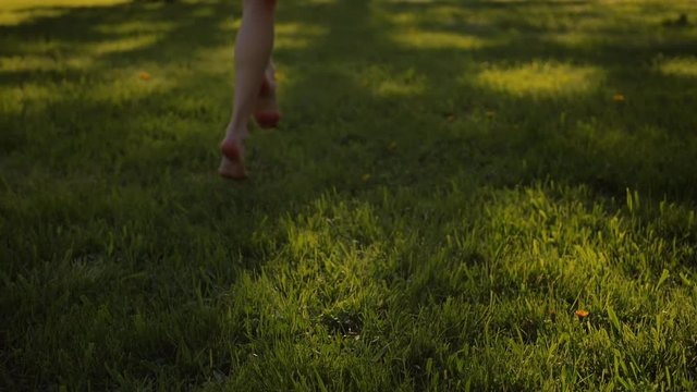 Lady's feet running by grass in park. No face. Sunset or surise. Real-time and slow motion mixed footage. Middle skirt, slender legs, barefoot.