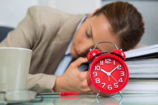 Woman asleep at office desk, alarm clock in foreground