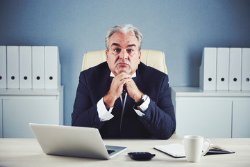 Calm wistful mature male in black business suit sitting propping chin with folded hands and looking...