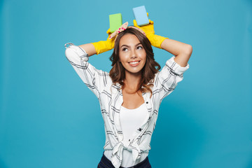 Photo of optimistic smiling housewife 20s in yellow rubber gloves for hands protection holding two sponges during cleaning, isolated over blue background