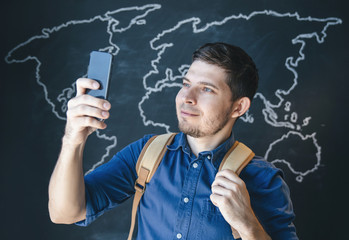 A guy in a blue shirt against a dark chalky wall with a smartphone and a backpack makes a photo.