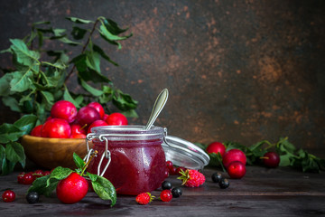 assortment of jam in glass jars, seasonal fresh berries and fruit plum, strawberry, currant, raspberries on a dark background selective focus with copy space, crop concept