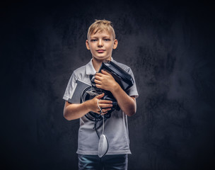 Blonde schoolboy dressed in a white t-shirt, holds full digital set for entertainment at a studio. Isolated on dark textured background.