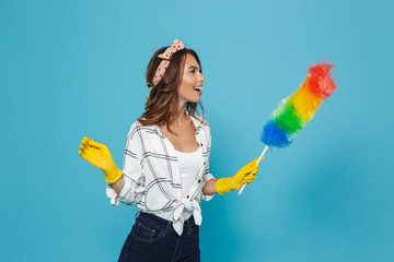 Photo of female houseworker 20s in yellow rubber gloves for hands protection holding colorful duster brush during dusting, isolated over blue background